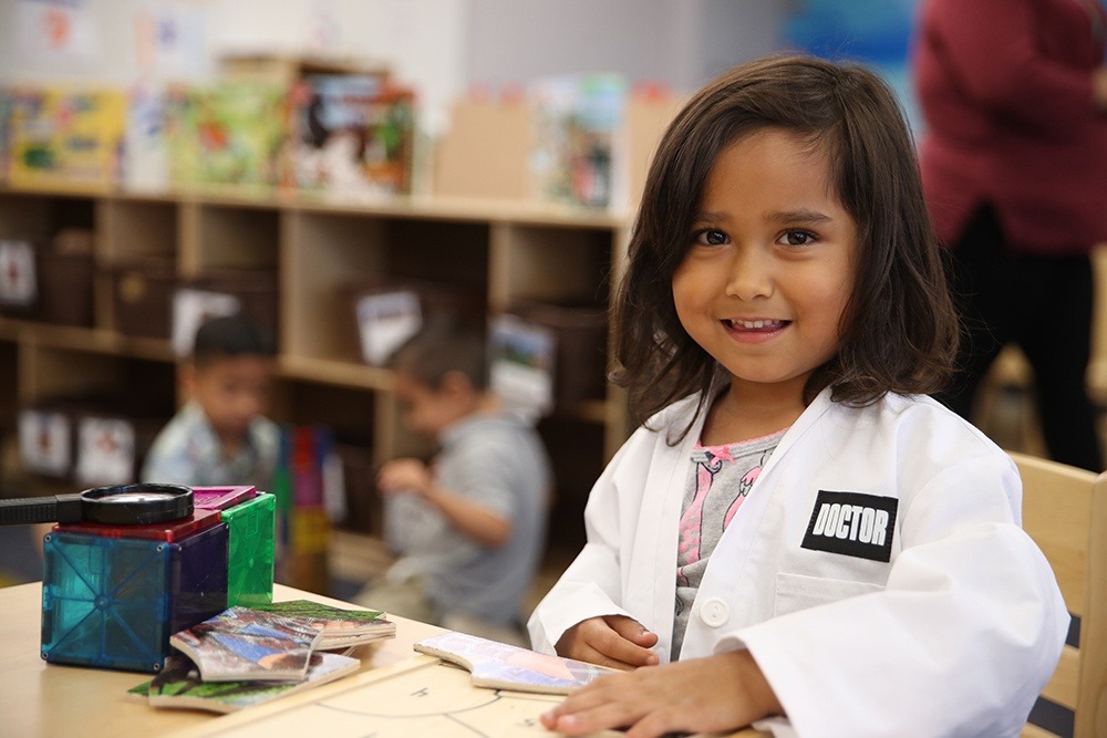 Young girl wearing a white lab coat labeled “Doctor” smiles while participating in a Think Together classroom activity.