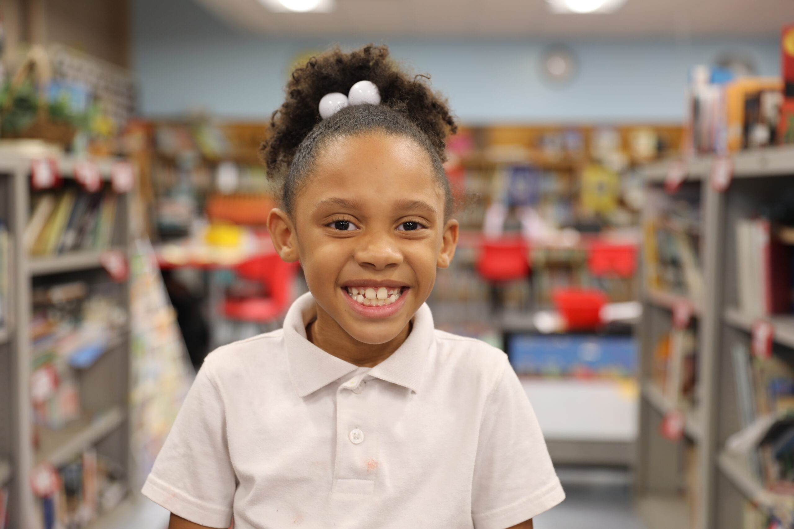 A smiling young girl wearing a white polo shirt stands in a colorful library filled with books and learning materials.