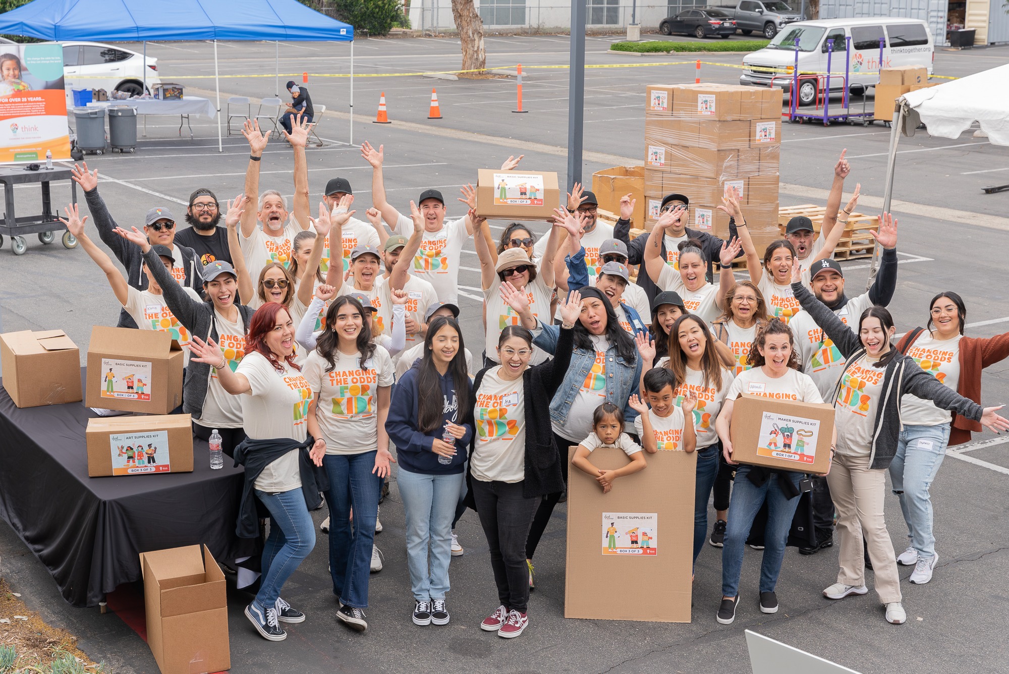 A large group of volunteers holding boxes and waving their hands while posing together in an outdoor parking area.