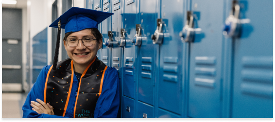 Student in blue cap and gown smiling beside school lockers on graduation day.